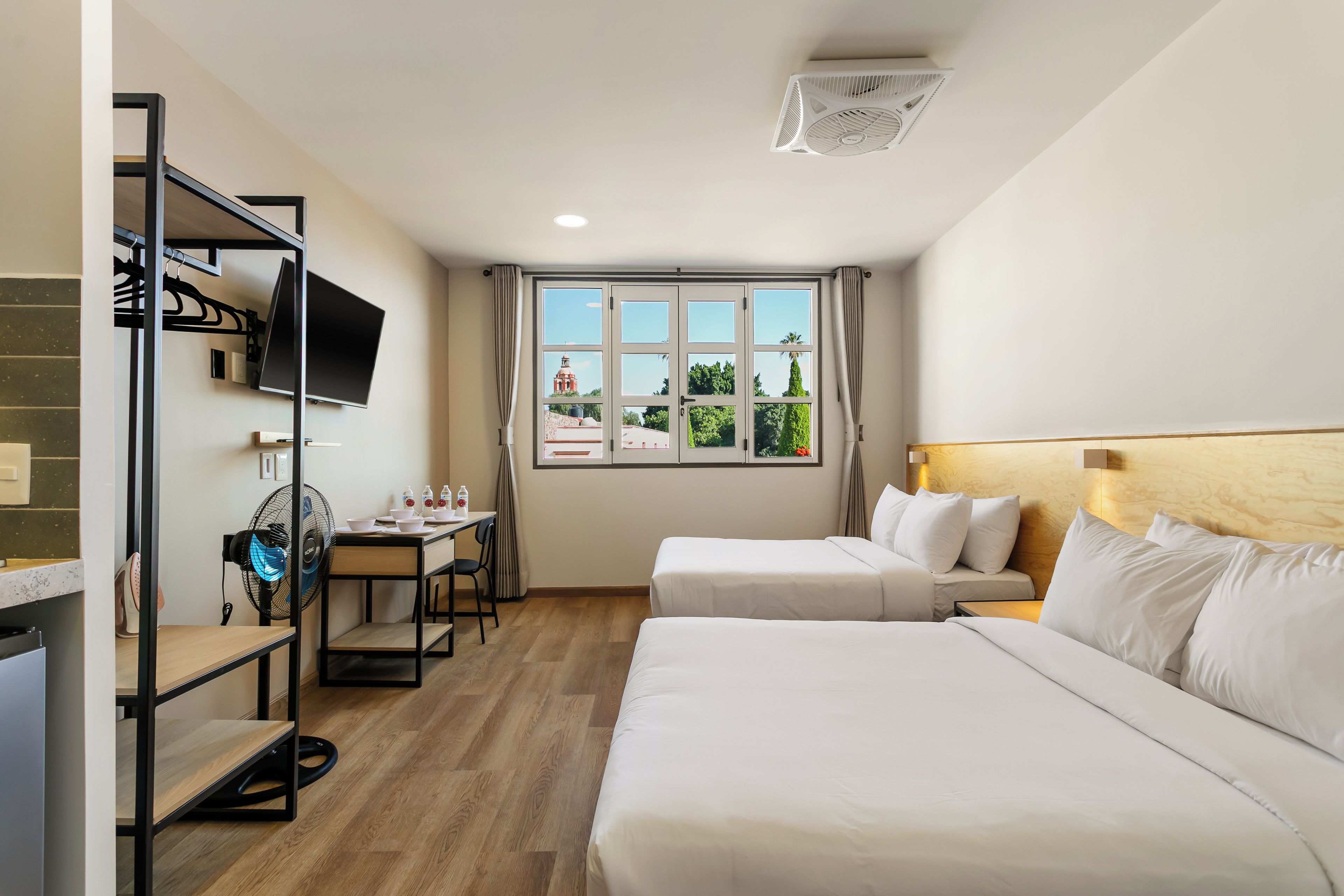 Ein Hotelzimmer mit zwei Betten, einem Schreibtisch mit Stuhl, einem Fernseher und einem Fenster mit Blick auf ein Gebäude und Bäume.