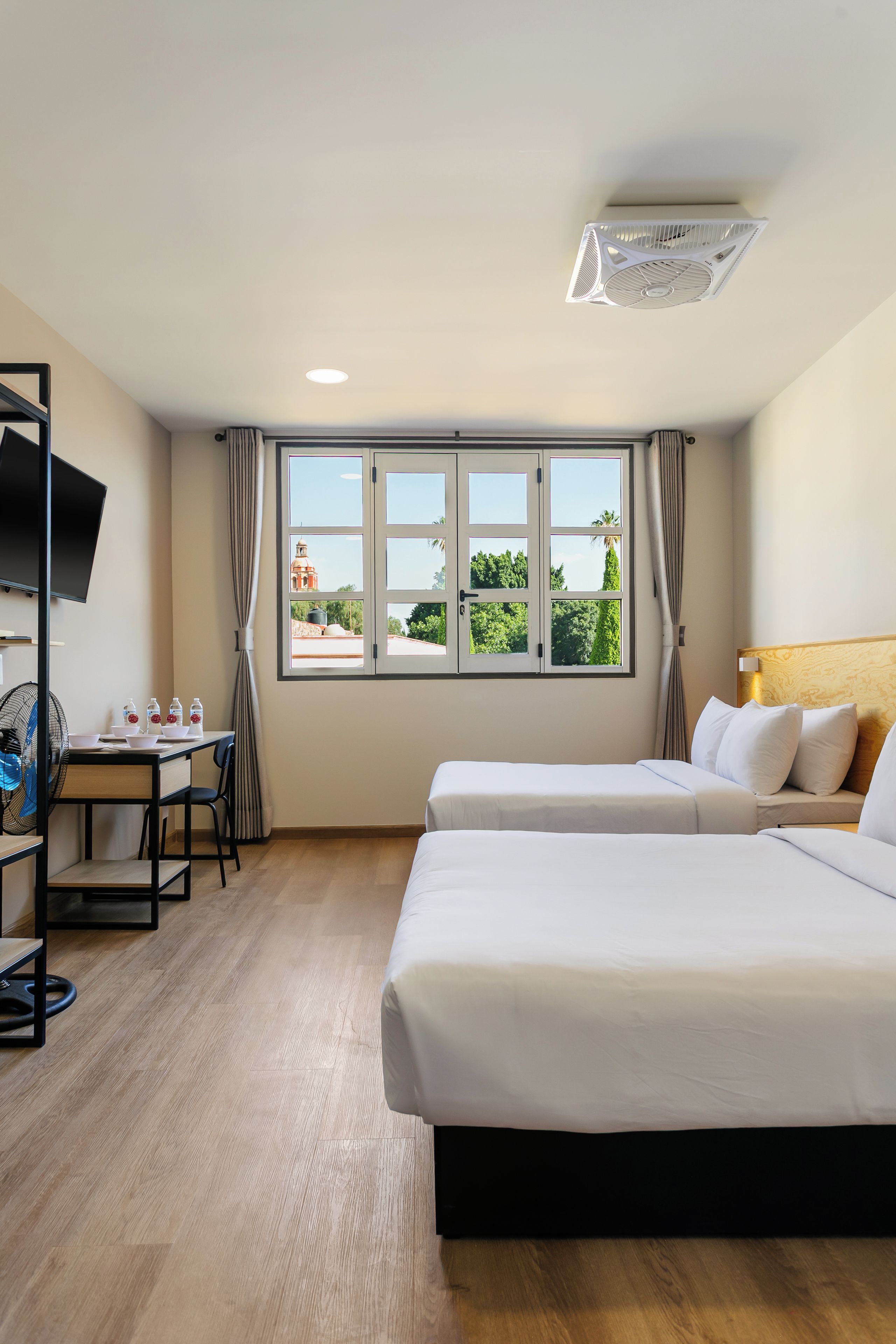 Habitación de hotel con dos camas, un escritorio, una silla, un televisor y una ventana con vistas a zonas verdes y un edificio.