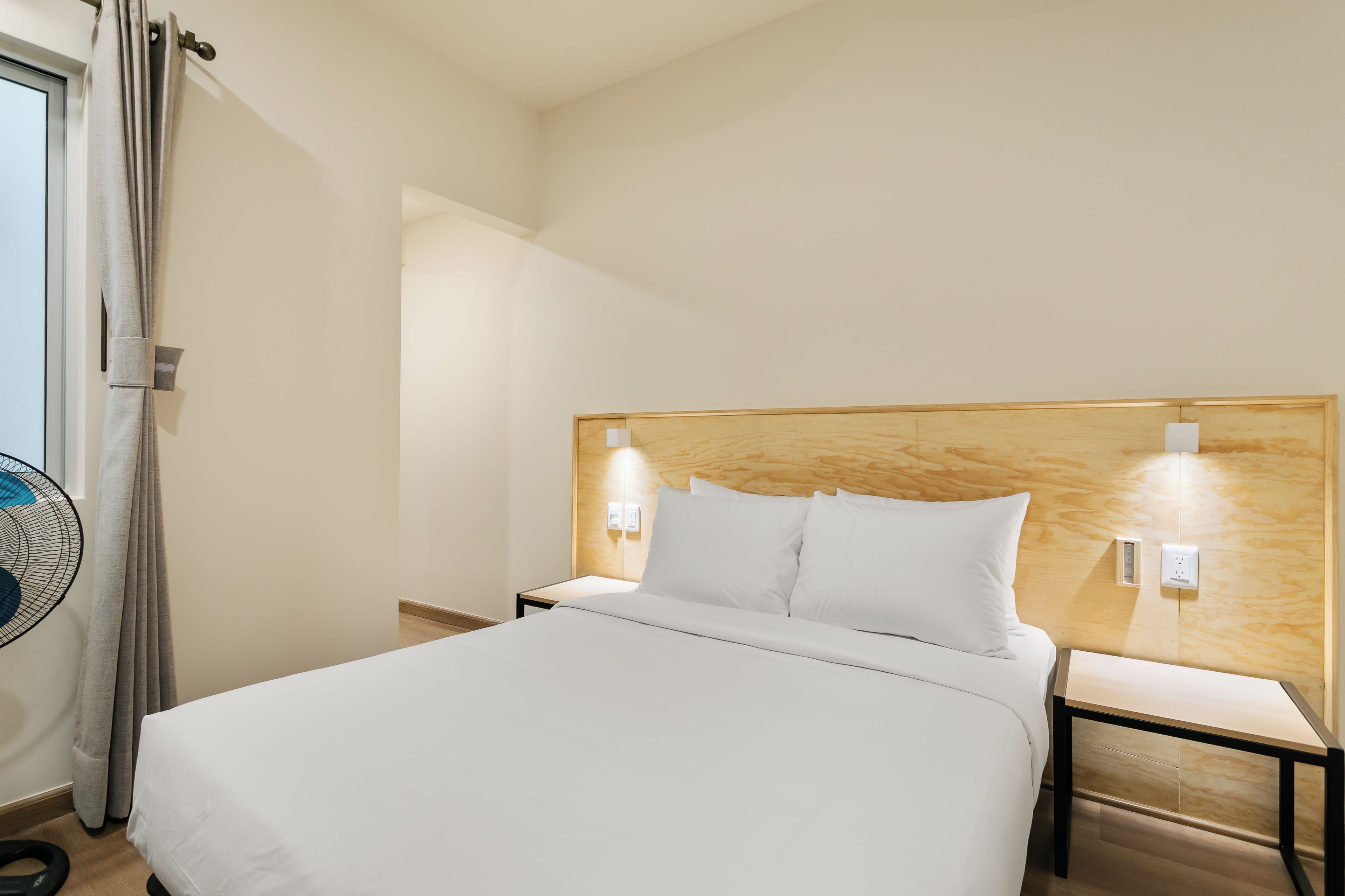 Une chambre d’hôtel comprenant un lit, des tables de chevet, une petite table, un ventilateur fixé au mur et une tête de lit en bois.