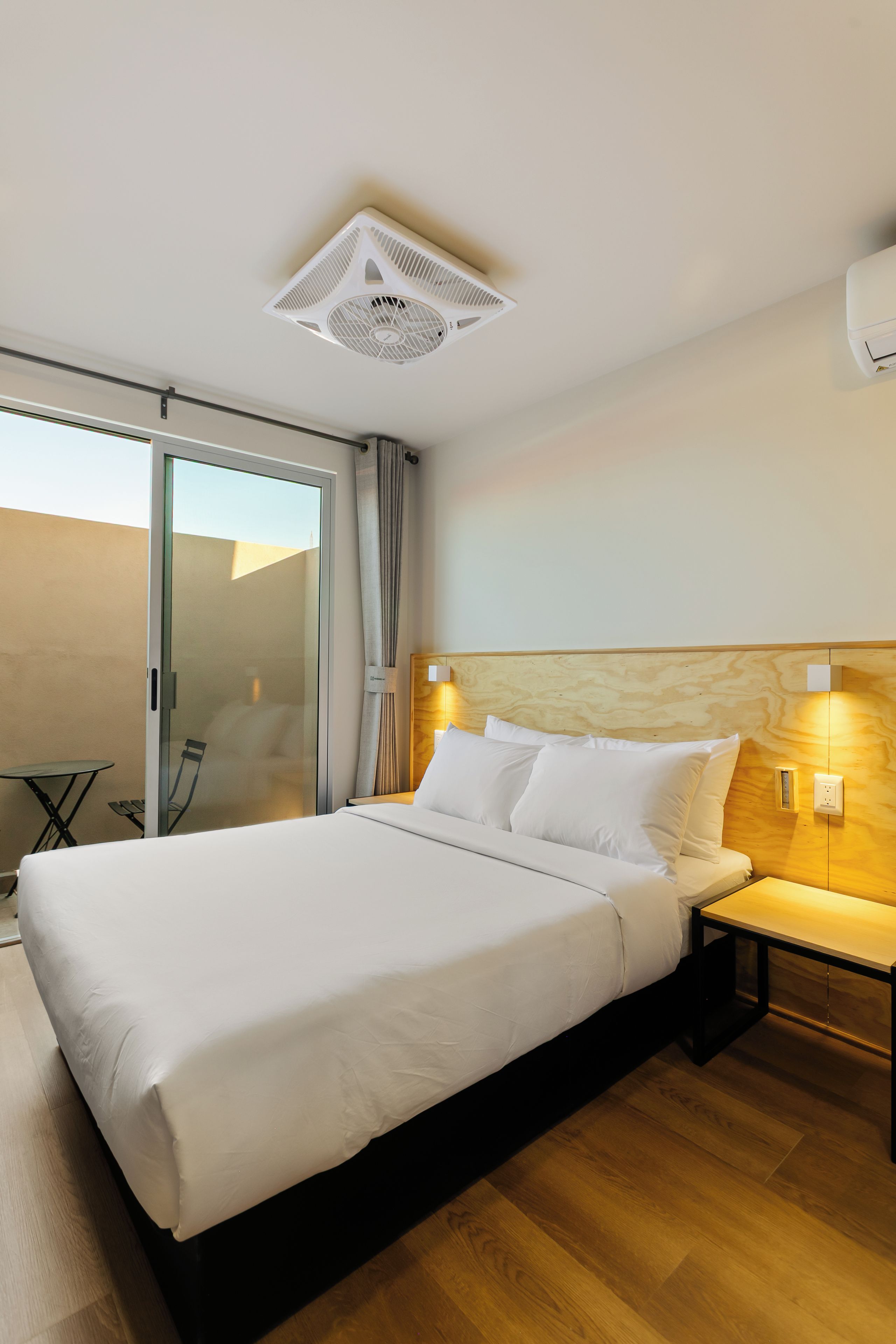 Habitació d'hotel amb un llit gran, una taula de nit, una làmpada penjada a la paret i una porta corredissa de vidre que dona a un balcó.