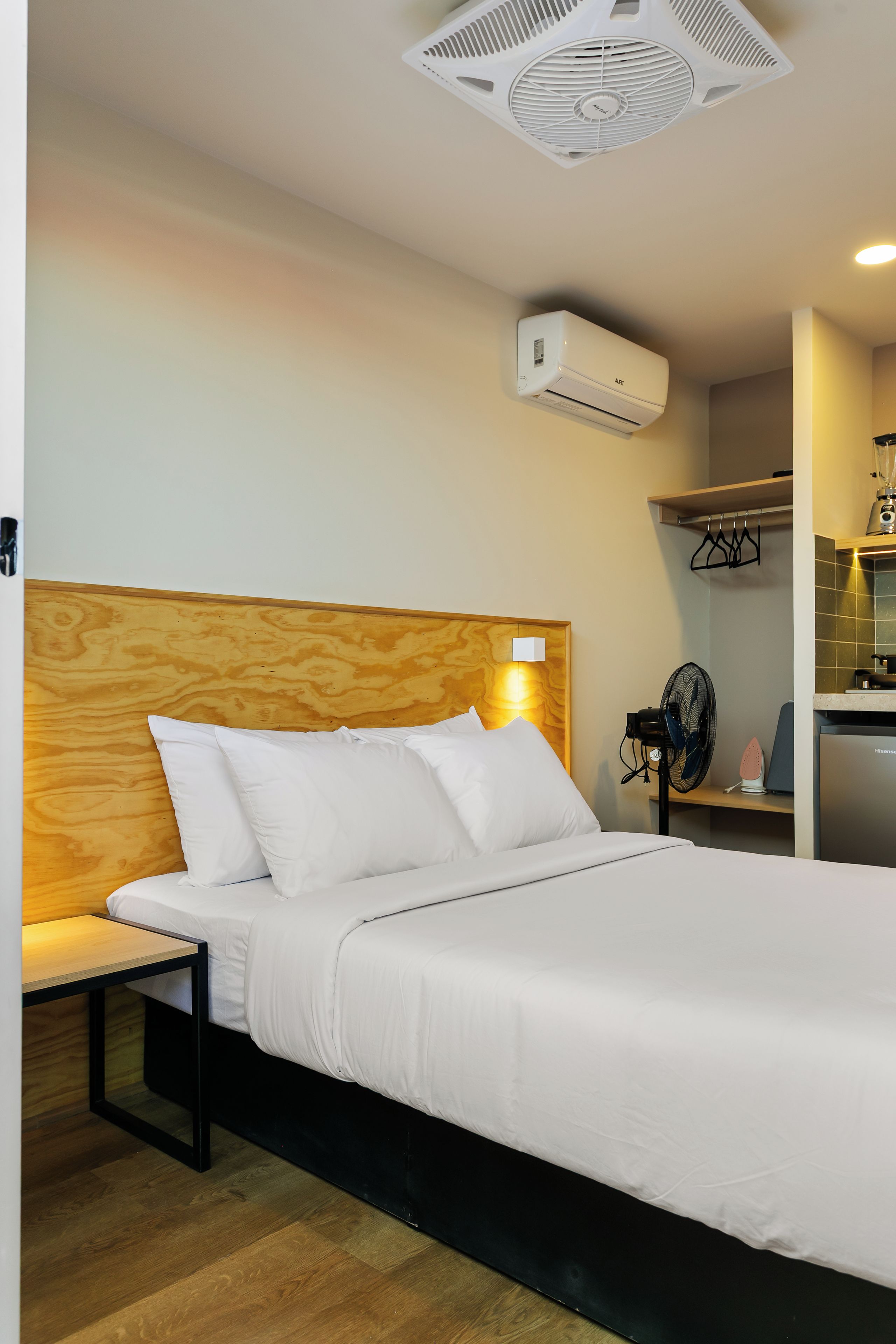 Une chambre d’hôtel avec une tête de lit en bois, un lit avec une literie blanche, une table de chevet, un climatiseur mural, un petit ventilateur et un coin cuisine équipé d’un mixeur et d’un grille-pain.