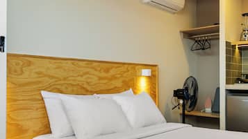 Habitación de hotel con cabecera de madera, cama con ropa de cama blanca, mesita de noche, aire acondicionado de pared, pequeño ventilador y área de kitchenette con licuadora y tostadora.