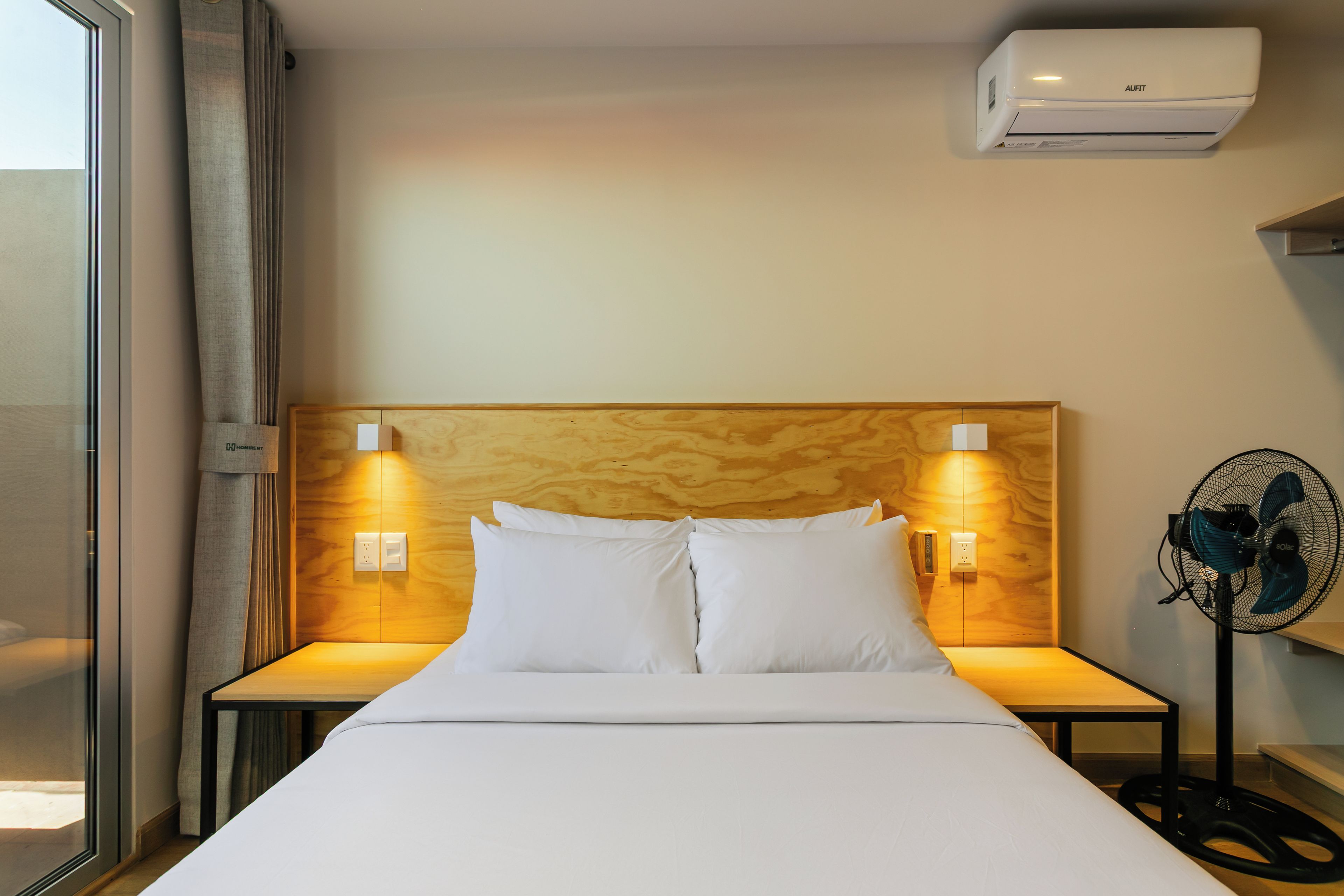 Een hotelkamer met een bed, twee bedlampen, een luchtverkoeler aan de muur en een staande ventilator.