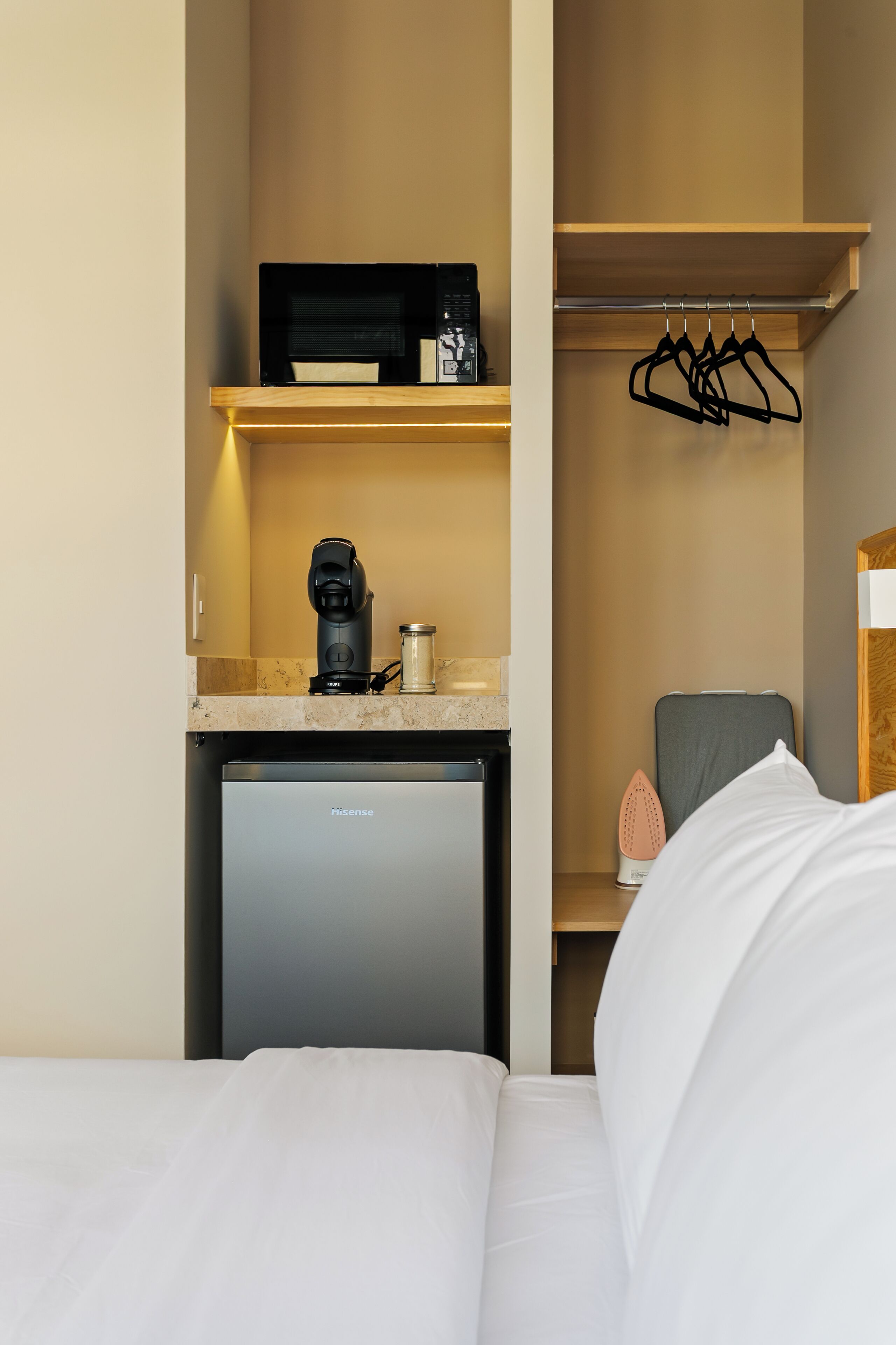 Una habitación de hotel compacta con cama, una kitchenette pequeña que incluye cafetera, un mini refrigerador y un estante de almacenamiento.