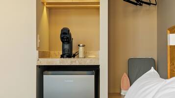 A compact hotel room with a bed, a small kitchenette with a coffee maker, a mini fridge, and a storage shelf.
