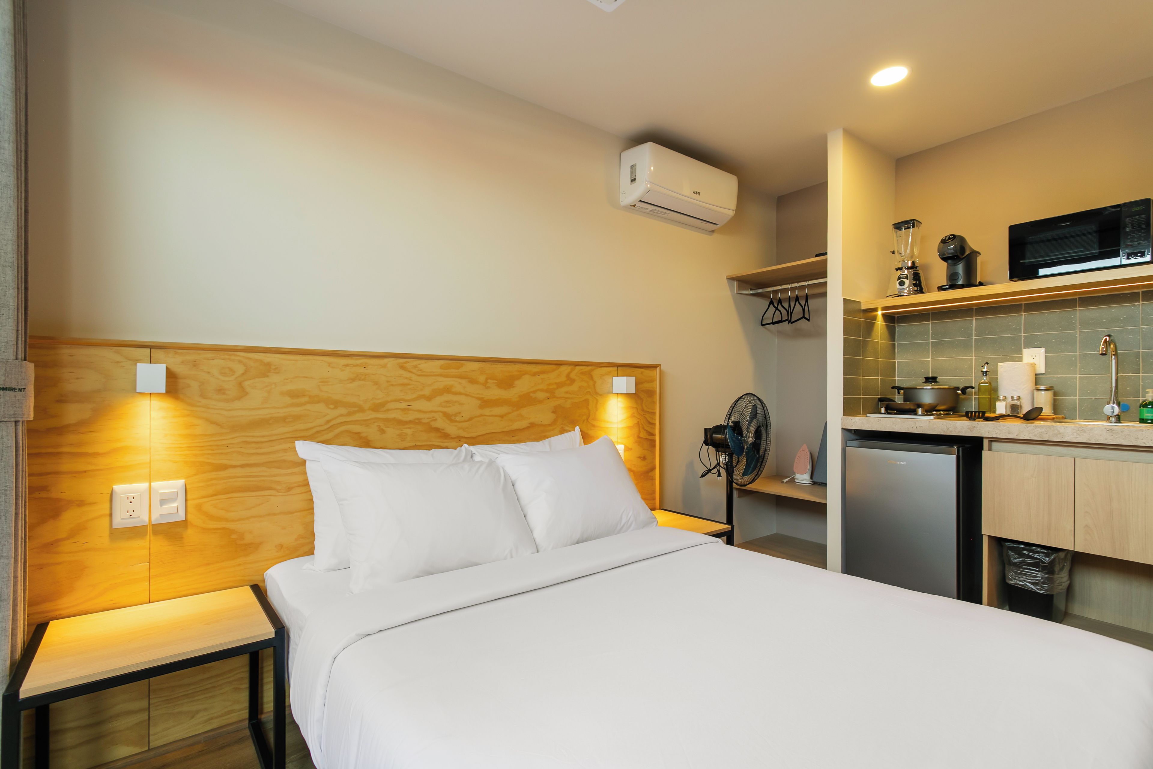 A hotel room with a bed, bedside table, wall-mounted air conditioner, and a kitchenette with a microwave, toaster, and sink.