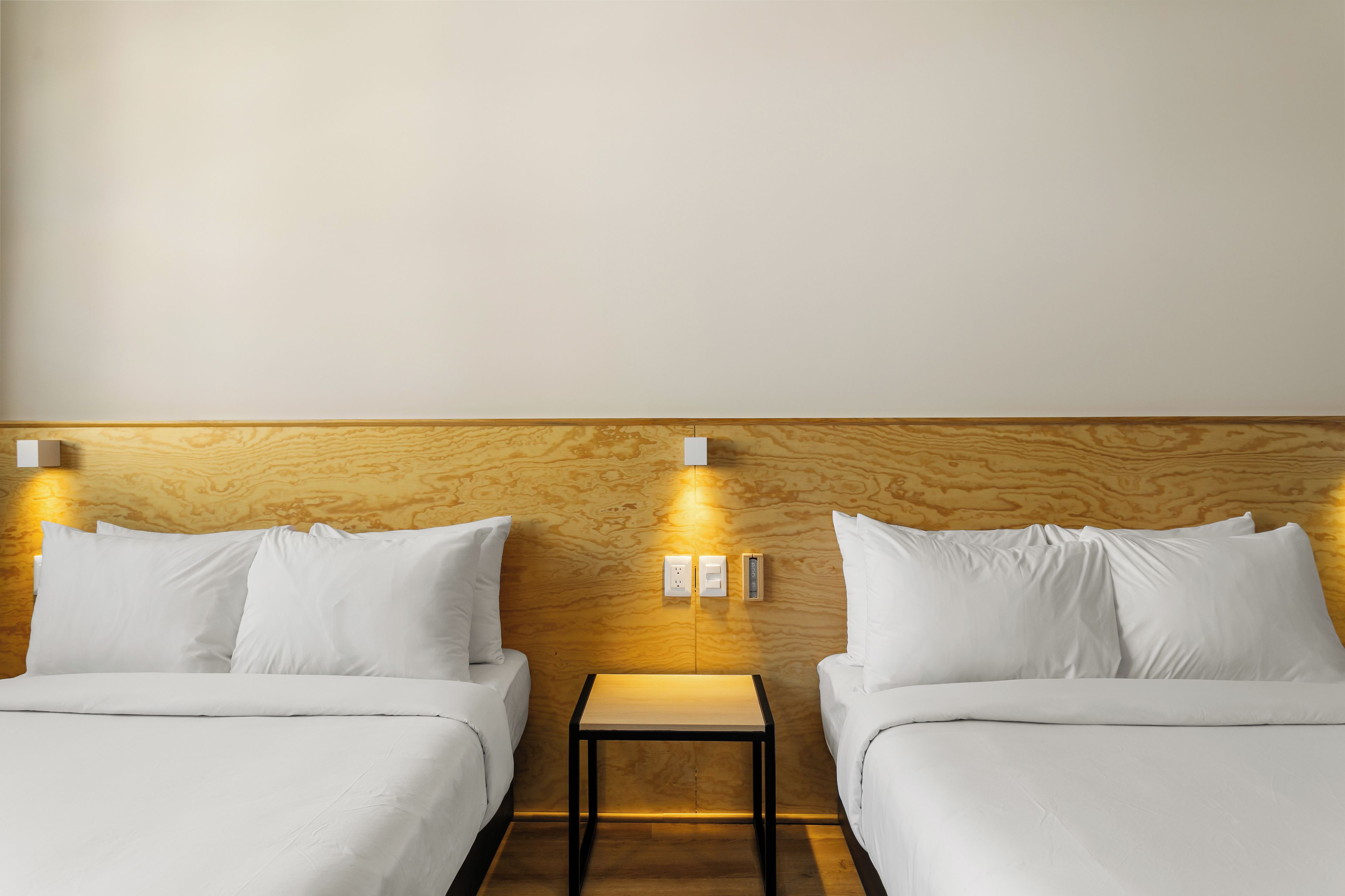 A hotel room with two beds, a wooden headboard, bedside tables, and wall-mounted light switches.