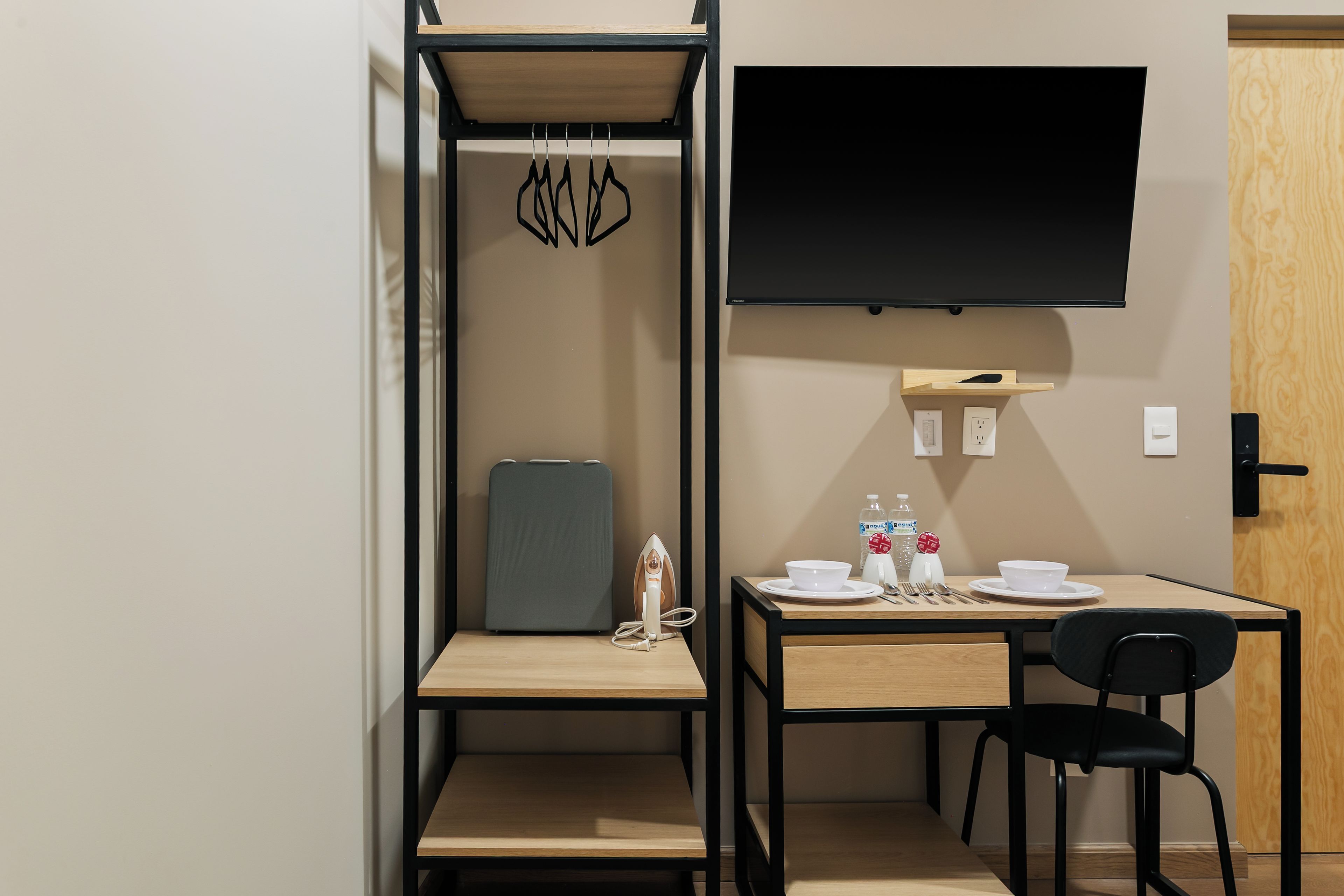 Une chambre de taille modeste, comprenant un lit, un bureau, une chaise, un téléviseur à écran plat et une petite étagère avec une bouteille et une tasse.