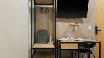 A compact room with a bed, a desk, a chair, a flat-screen TV, and a small shelf with a bottle and a cup.