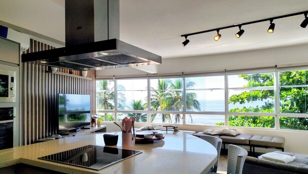 A modern kitchen with a central island, a dining area with a sofa, and large windows with a view of greenery and the sea. - Condomínio Praias do Guarujá (Guarujá)