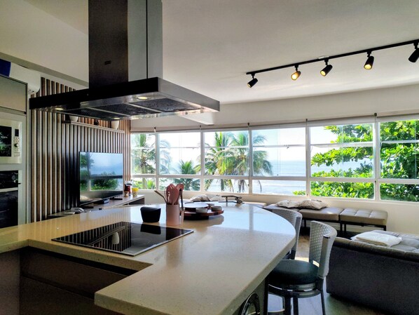 A modern kitchen with a central island, a dining area with a sofa, and large windows with a view of greenery and the sea.