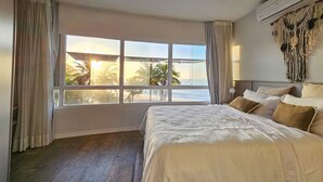 A bedroom with a large window, a bed with white and brown bedding, and a macrame wall decoration. - Condomínio Praias do Guarujá (Guarujá)