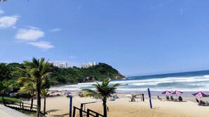 A beach with palm trees, beach umbrellas, and a volleyball net. - Condomínio Praias do Guarujá (Guarujá)
