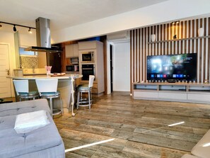 Una sala de estar moderna con un sofá gris, un televisor sobre un mueble de madera y una zona de cocina con microondas y horno.