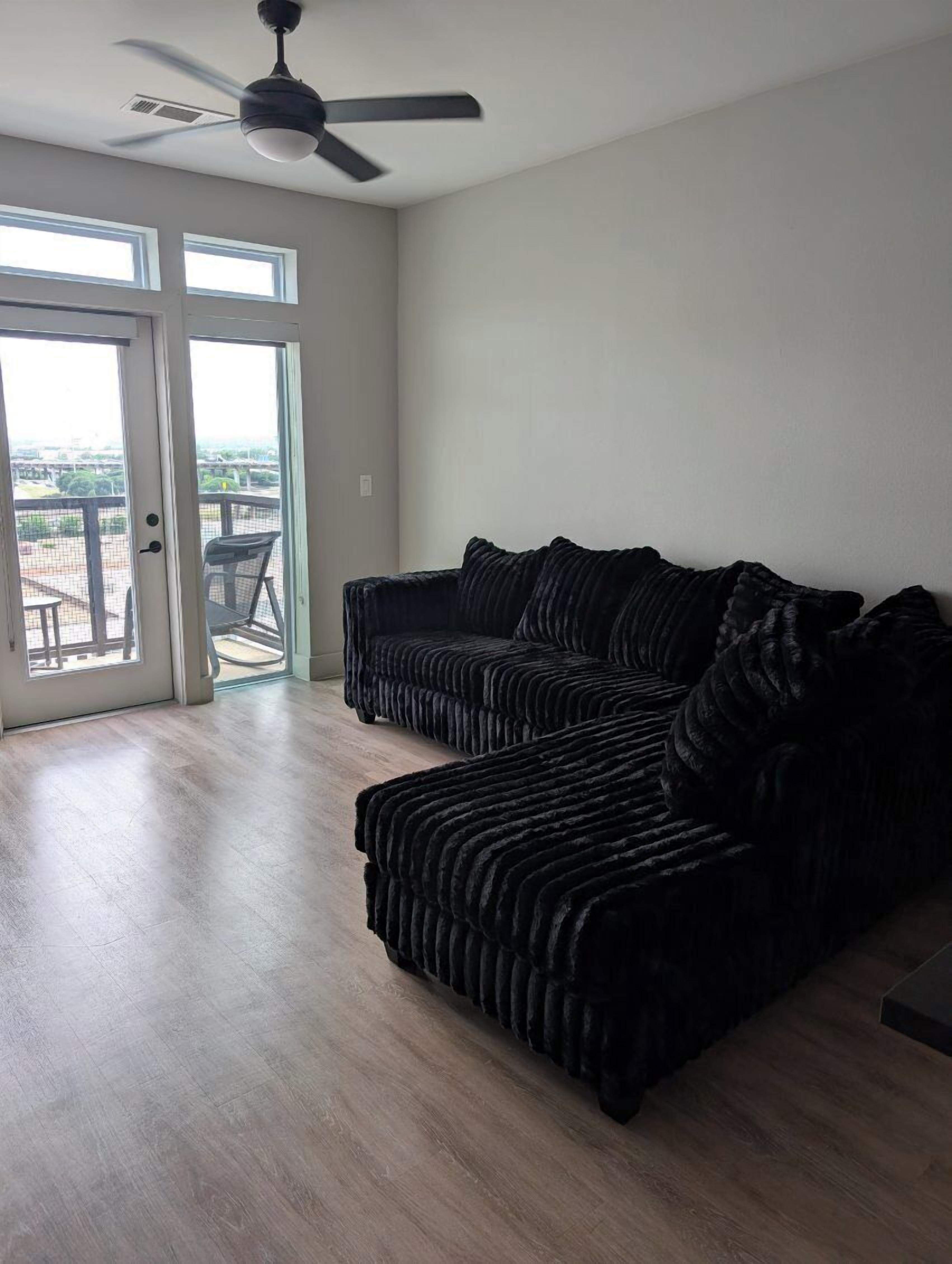 A spacious living room with a dark-colored sectional sofa, a ceiling fan, and a sliding glass door leading to an outdoor patio.