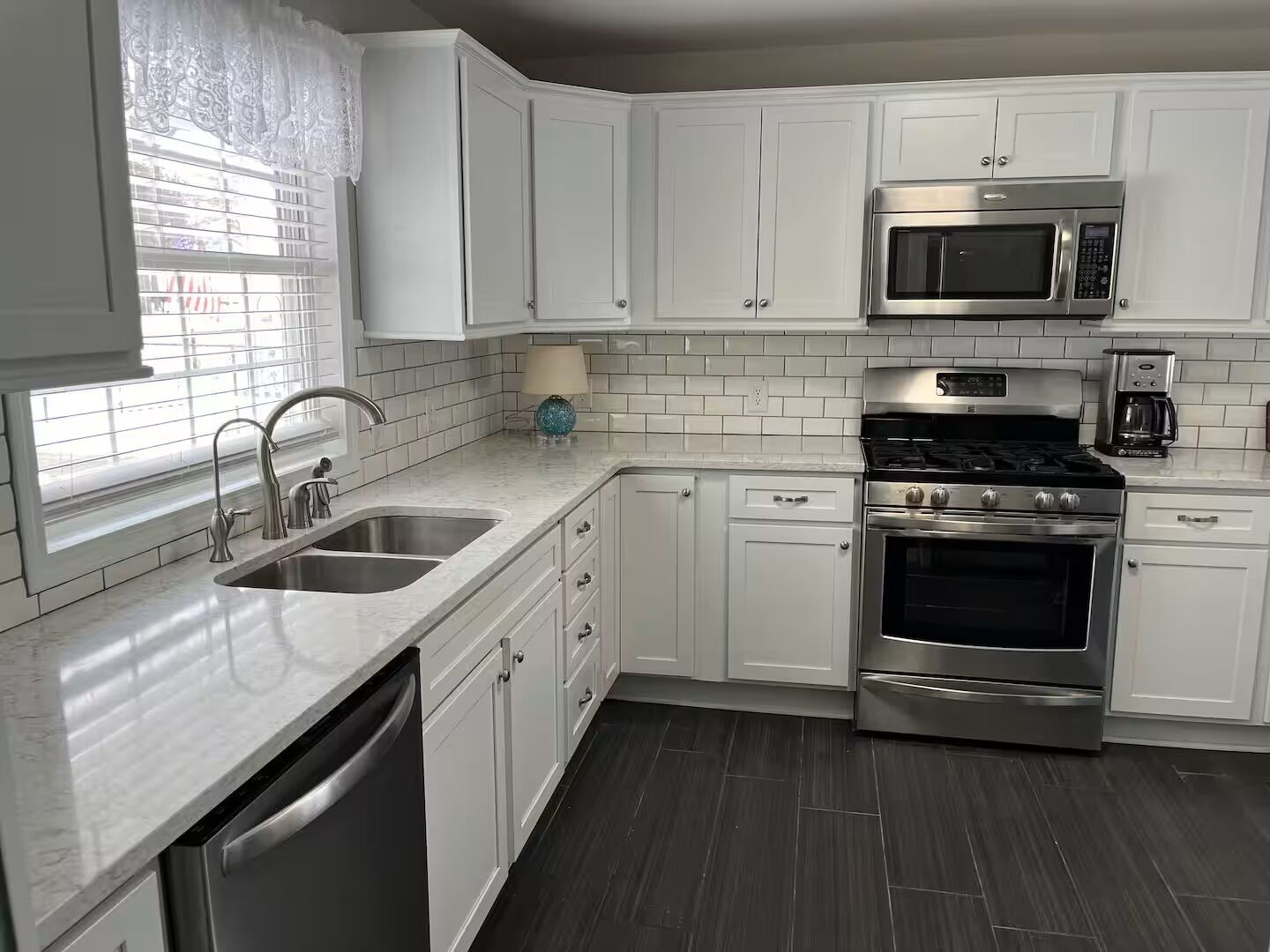 A modern kitchen with white cabinets, stainless steel appliances, a dishwasher, and a window with blinds.