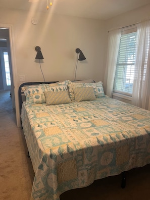 A bedroom with a bed, two wall-mounted lamps, a window with blinds, and a door leading to another room.