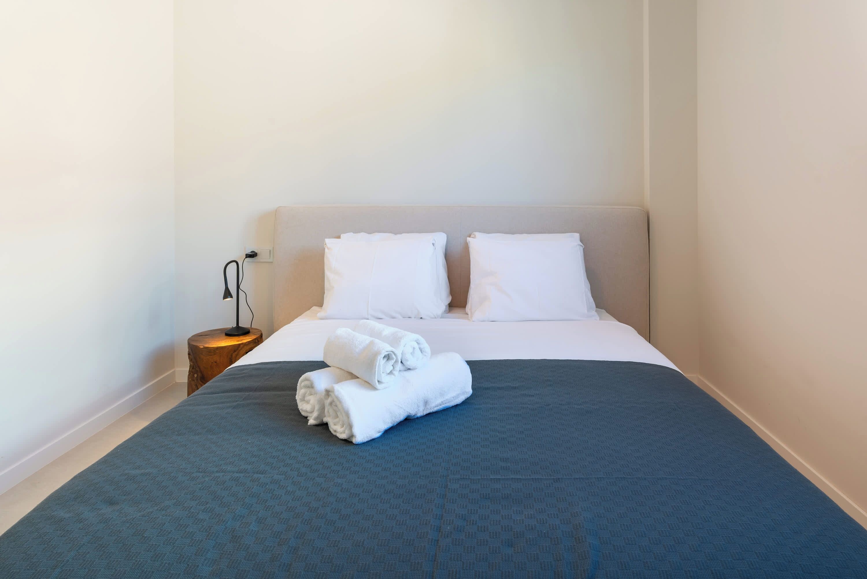 Een net opgemaakt bed met witte lakens en een grijze deken, een bedlampje en een houten nachtkastje.