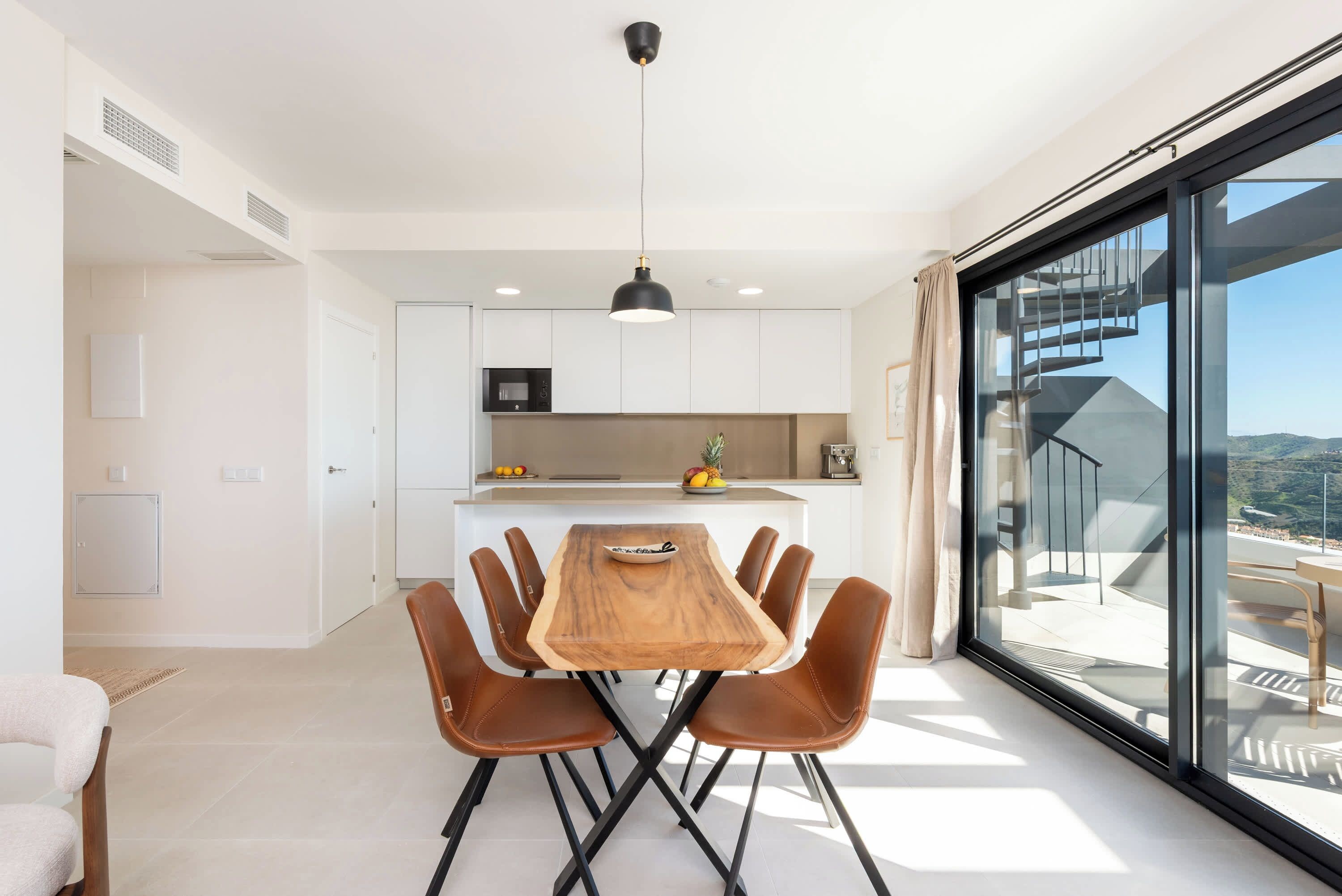 A modern kitchen with white cabinets, a wooden dining table with six chairs, and a large window offering a view of a staircase and landscape.