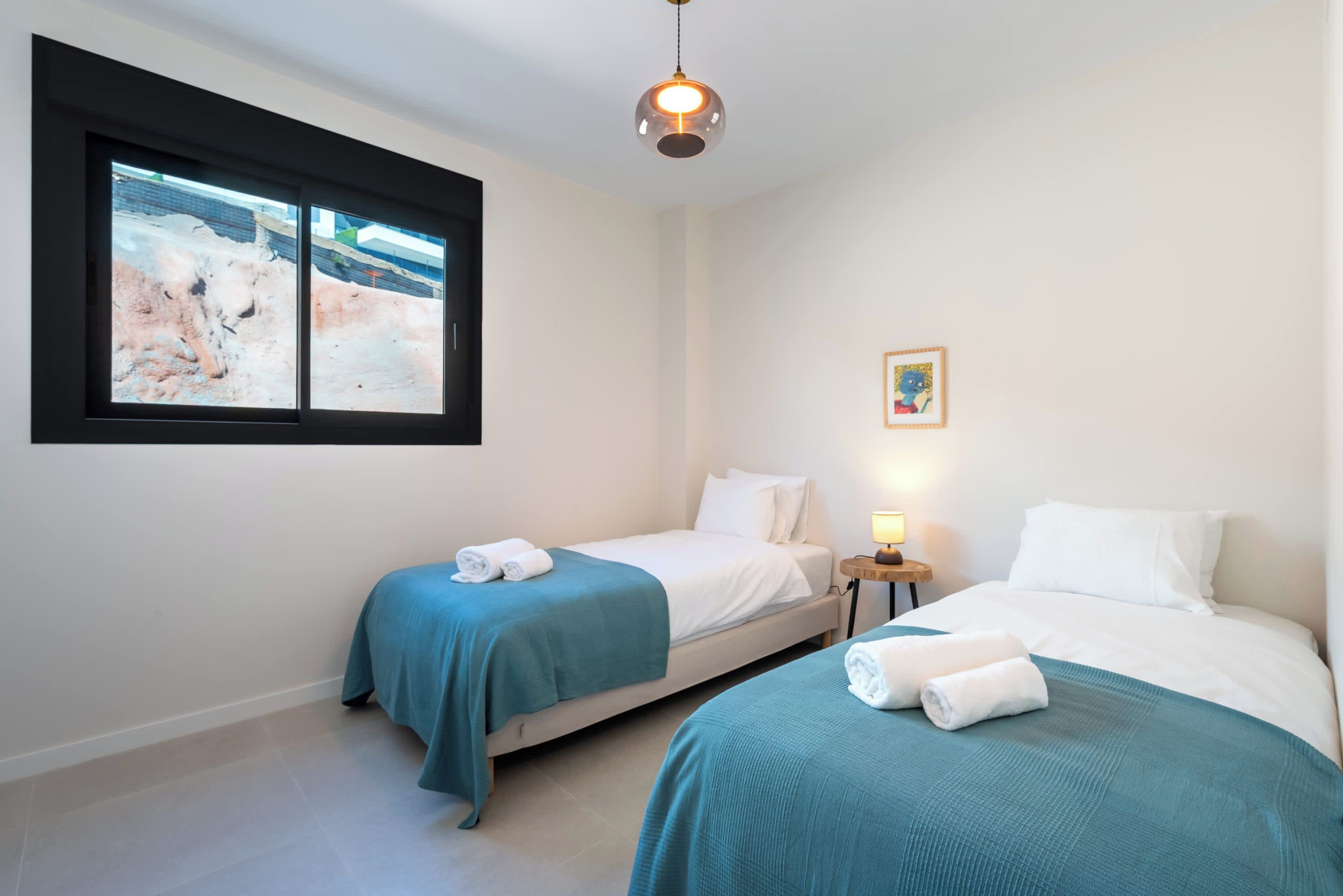 Hotelkamer met twee bedden, een raam met uitzicht op een bouwplaats, een hanglamp en een klein nachtkastje met een lamp.