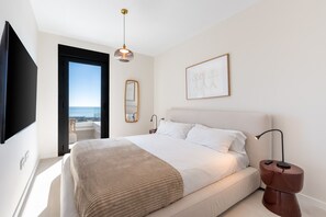 A modern bedroom with a large bed, a bedside table with a lamp, a mounted TV, a framed artwork, and a view of the sea through an open door. - Bali II | Costa del Sol Penthouse with Amazing Views (Rincón de la Victoria)