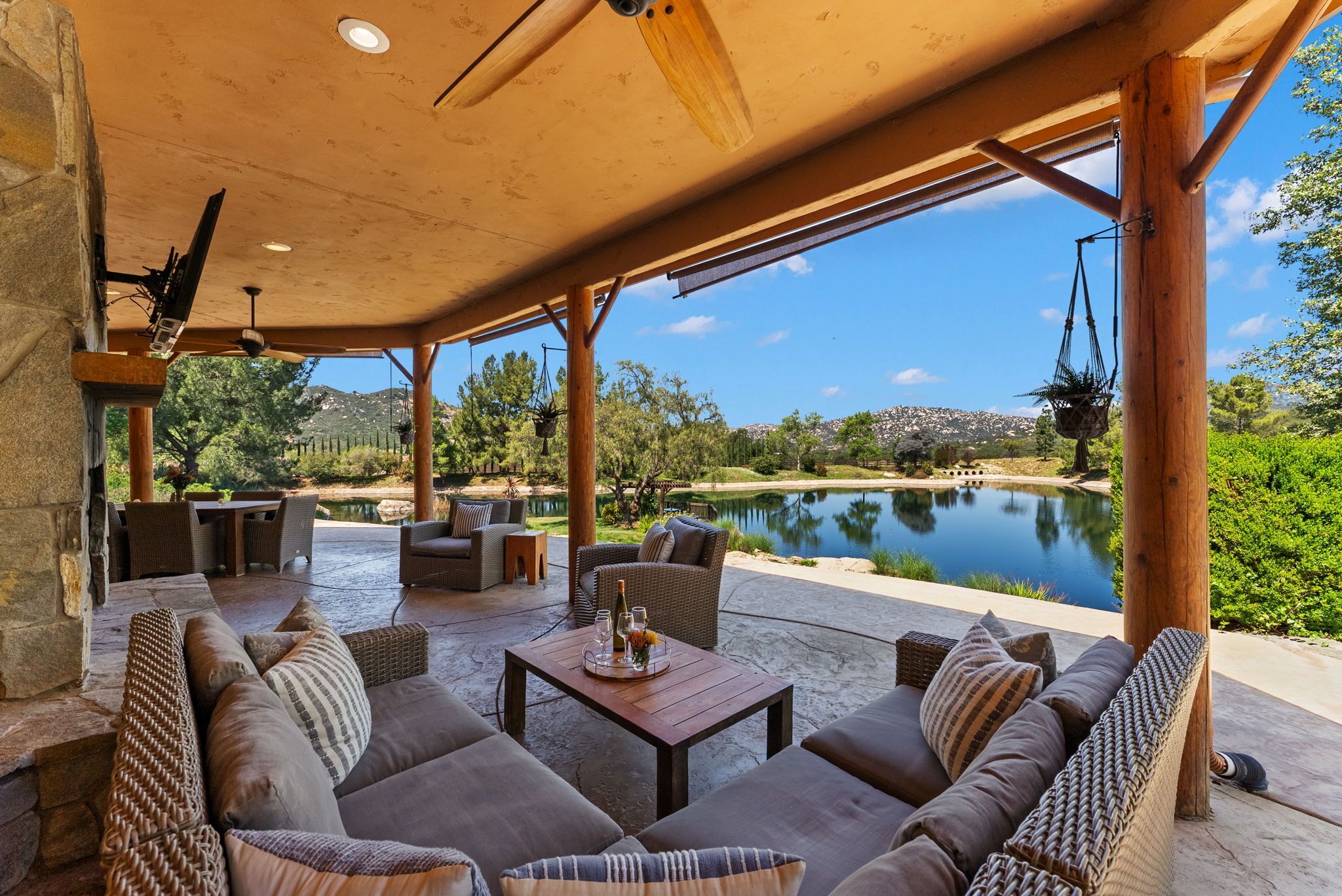 An outdoor patio with wicker furniture, a table with glasses and a bottle, and a view of a lake and hills.