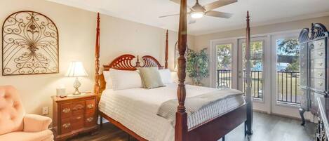 A four-poster bed with a wooden headboard, a nightstand with a lamp, a pink armchair, and a large window with a view of the outdoors.