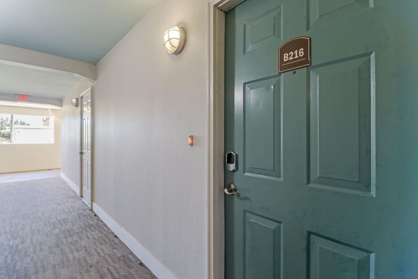 Corredor com uma porta verde identificada como B216, luminária e um interfone.