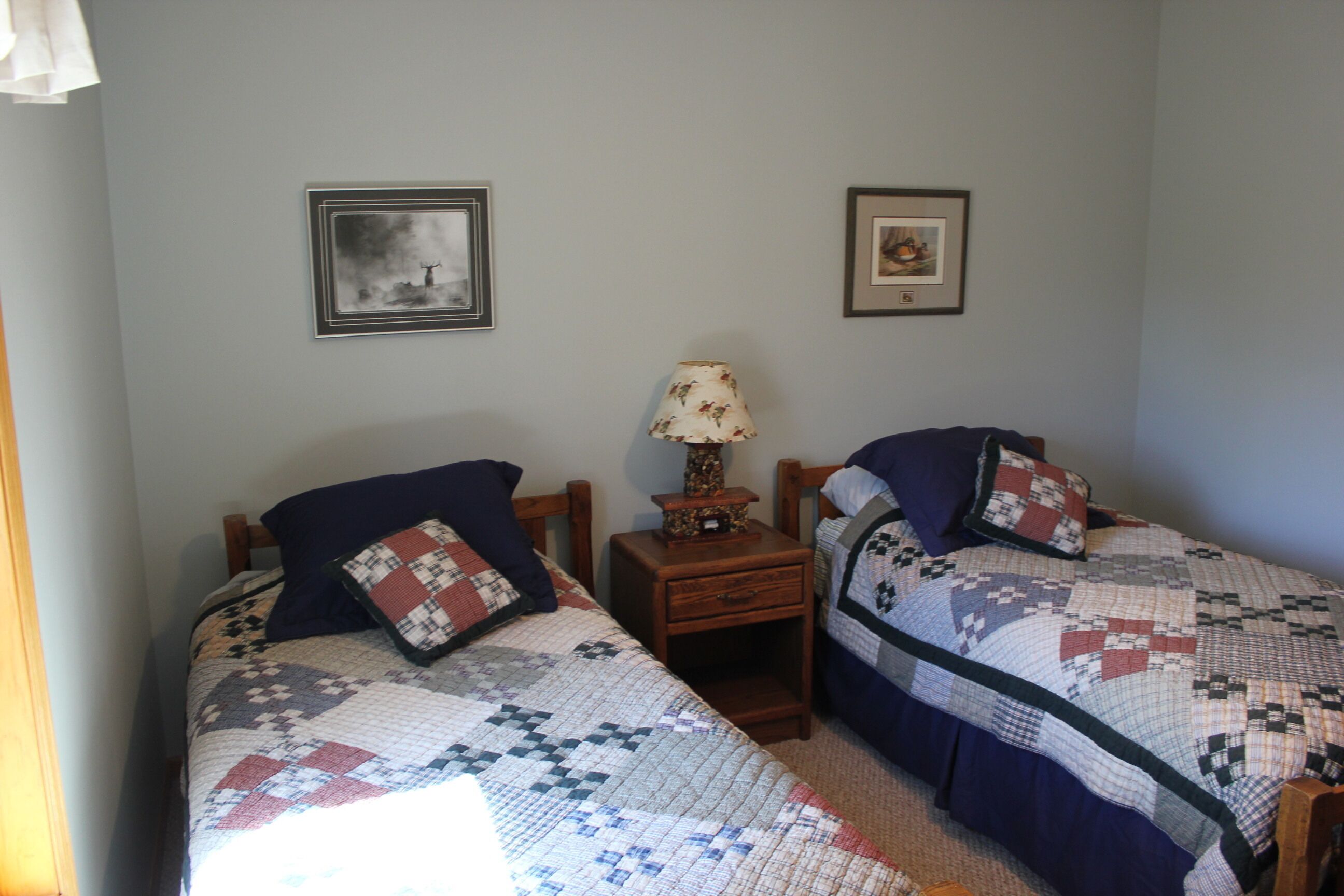Un dormitorio con dos camas individuales, una mesita de noche con lámpara y dos cuadros enmarcados en la pared.