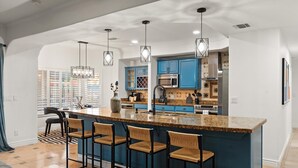 Een moderne keuken met blauwe kastjes, een granieten eiland met barstoelen, hanglampen en uitzicht op buiten.