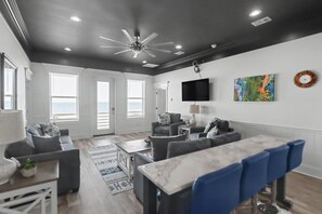 A modern living room with a ceiling fan, a flat-screen TV, and a dining area with bar stools. - The Gulf Pearl - Beach House (Long Beach)
