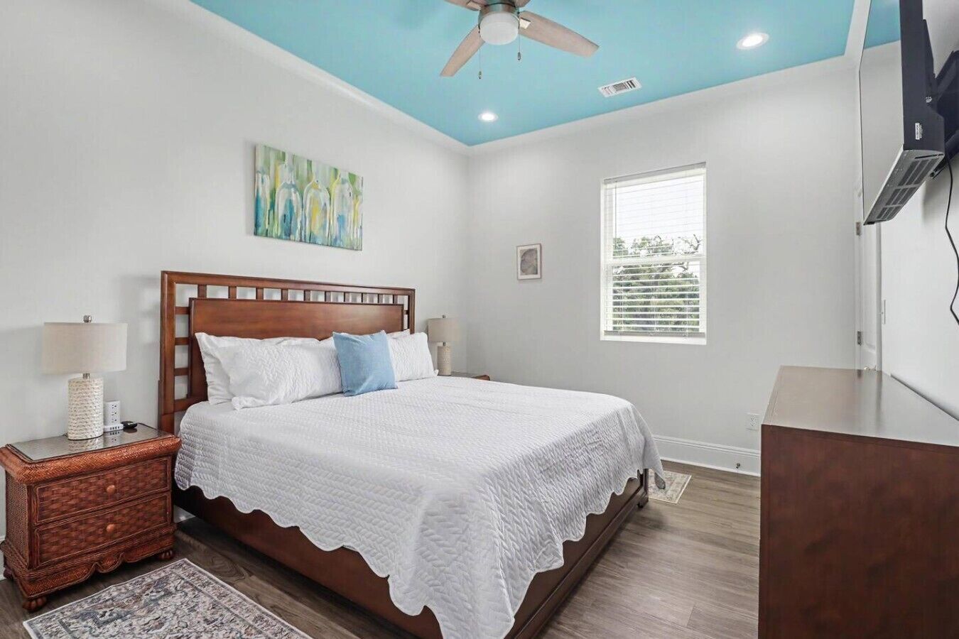A bedroom with a wooden bed, a nightstand, a lamp, a painting, a window, and a ceiling fan.
