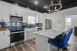 A modern kitchen with white cabinets, a central island, blue bar stools, and a chandelier. - The Gulf Pearl - Beach House (Long Beach)
