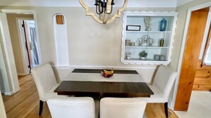 A dining area with a dark wooden table, white cushioned chairs, a chandelier, and a decorative shelf with glassware. - Stylish 2BR + Office | Near Cleveland Clinic+Shops (South Euclid)