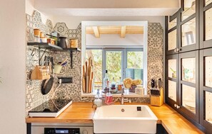 Una cocina con un lavamanos blanco, encimeras de madera, utensilios colgando y una ventana con vistas a árboles.
