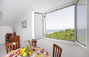 Ein Essbereich mit Tisch für zwei Personen, Obstschale und Blick auf eine Küstenlandschaft durch ein großes Fenster.