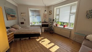 Ett sovrum med en säng, byrå, skrivbord, element, krukväxter och stora fönster.