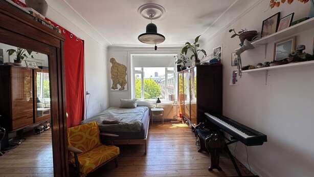 Ett rum med en säng, en soffa, ett piano, en spegel och ett fönster med utsikt över byggnader.