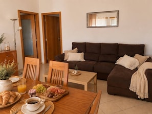 Ein gemütlicher Wohnbereich mit braunem Sofa, Esstisch aus Holz und reichhaltigem Frühstücksbuffet mit Croissants, Früchten und Kaffee.