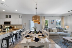 Eine moderne Küche mit weißen Schränken, ein Essbereich mit einem weißen Tisch, gedeckt für vier Personen, und ein graues Sofa im Wohnzimmer.
