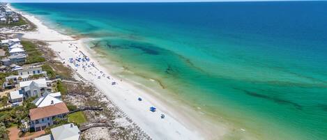Luchtbeeld van een strand met helder turquoise water, een zandstrand en strandwoningen met zichtbare daken en ramen.