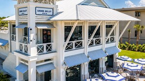 Um edifício costeiro com uma placa indicando 'SHUNK GULLEY OYSTER BAR', mesas ao ar livre com guarda-sóis e um céu claro.