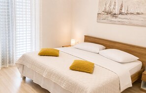 A neatly made bed with white linens and two yellow pillows, a wooden headboard, a bedside table with a lamp, and a painting of sailboats on the wall. - Stunning apartment in Tribanj Sibuljina with WiFi (Tribanj Sibuljina)