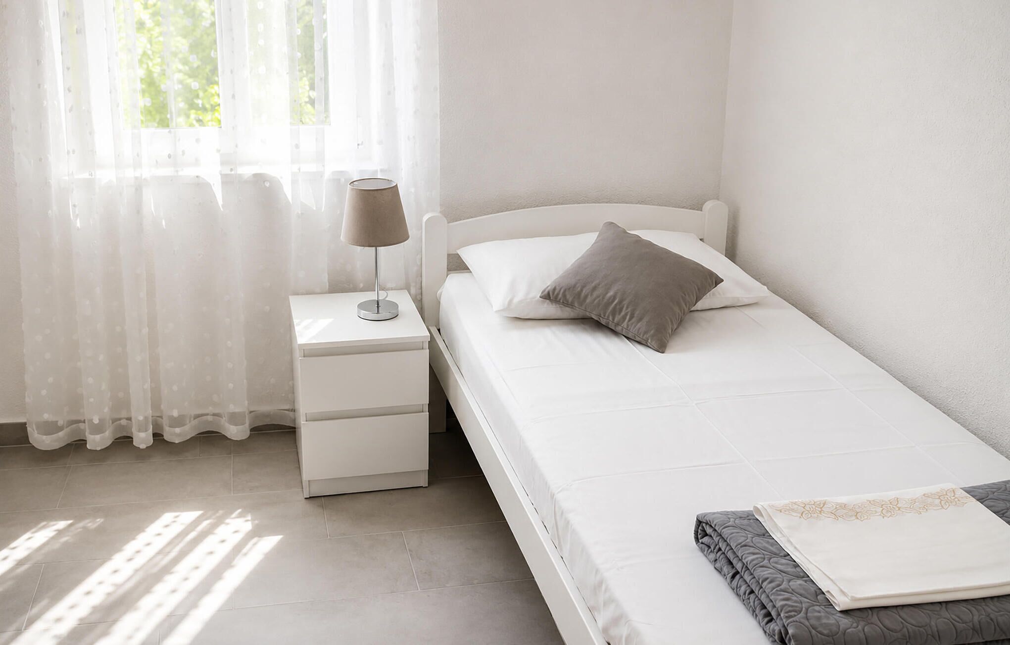 A single bed with a pillow, a bedside table with a lamp, and a window with sheer curtains.