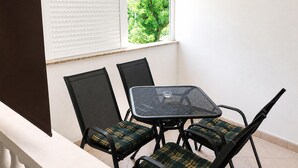 Un balcón con mesa y sillas, una ventana con persianas, y vistas a la vegetación.