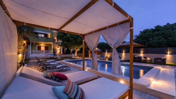 A poolside area with sun loungers, a covered seating area, and a view of a building in the background.