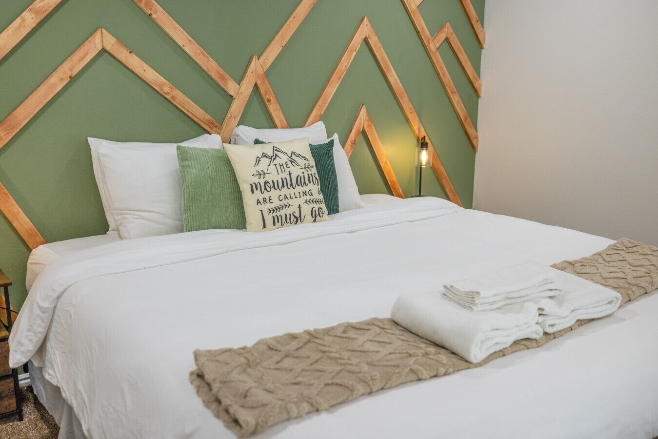 A neatly made bed with white bedding and a beige throw blanket, a pillow with a mountain-themed design, and a wooden headboard with a geometric pattern.