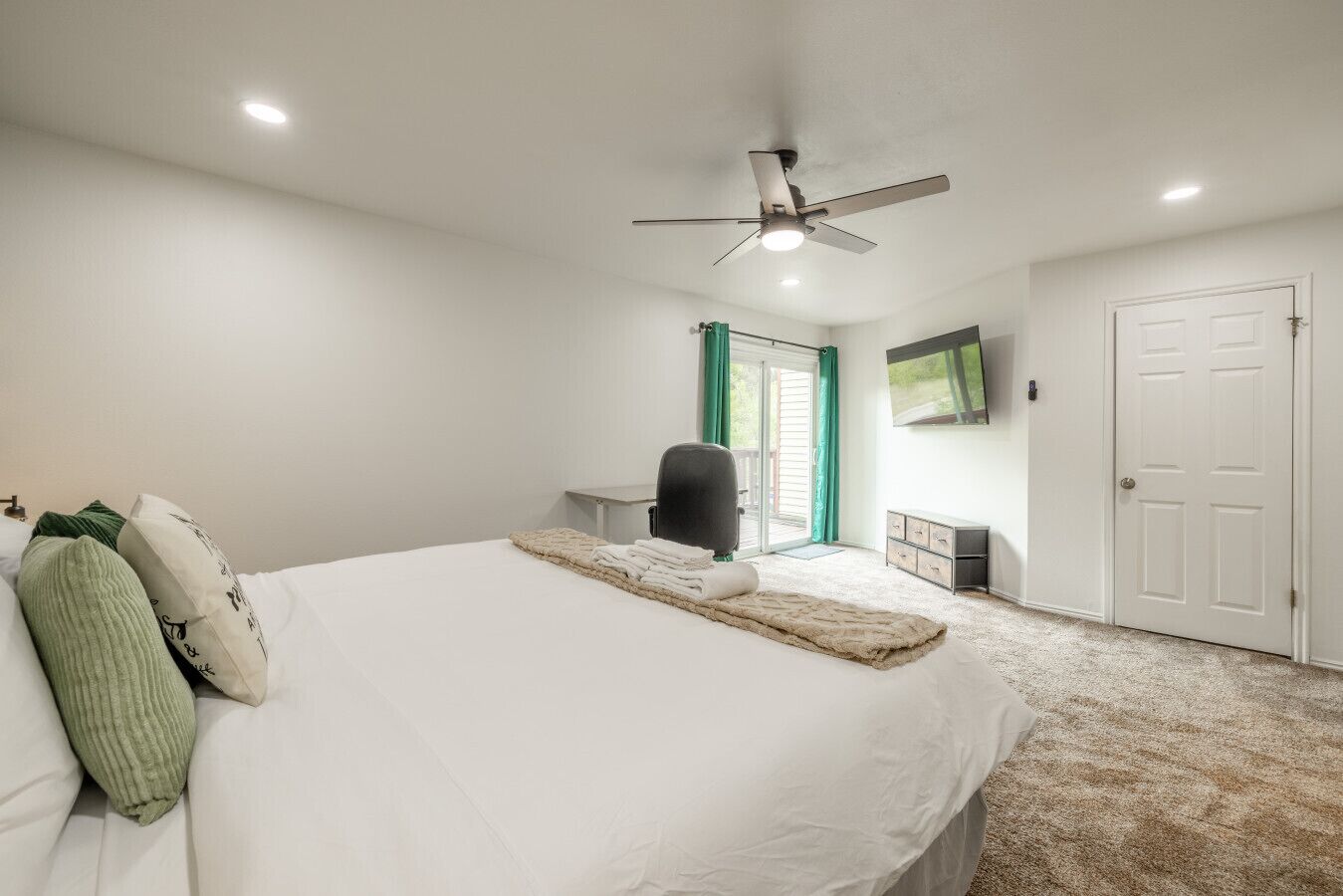 A bedroom with a bed, a desk, a chair, a ceiling fan, a TV, and a door.