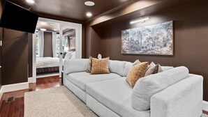 Un salon moderne avec un canapé d’angle gris, un grand tableau abstrait, des spots encastrés au plafond et un téléviseur à écran plat fixé au mur.
