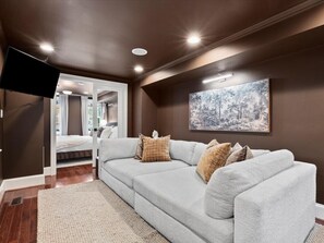 Ein modernes Wohnzimmer mit einer grauen Eckcouch, einem großen abstrakten Gemälde, deckenmontierten Leuchten und einem flachbildfernseher an der Wand.